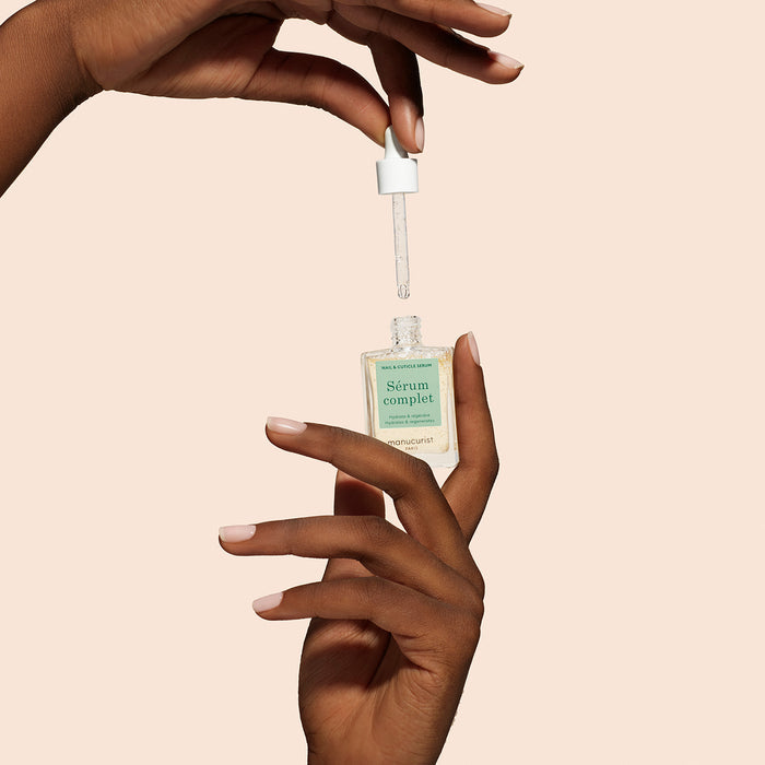 A hand holds a glass bottle of Manucurist Nail Hydrating Serum as another hand dispenses the treatment with a dropper against a soft beige background.
