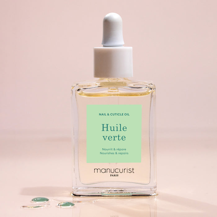 A glass dropper bottle of Manucurist Nourishing Cuticle Oil sits on a light surface with a few oil drops nearby. The pale green label with white and dark green text suggests enhanced nail hydration.