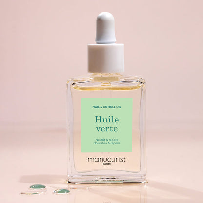 A glass dropper bottle of Manucurist Nourishing Cuticle Oil sits on a light surface with a few oil drops nearby. The pale green label with white and dark green text suggests enhanced nail hydration.