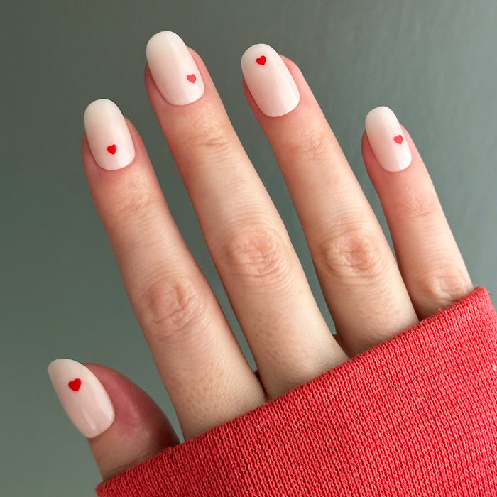Hand wearing Red Hearts | Short | Round - Tab Press-On nails with neutral base and tiny red heart accents against gray background
