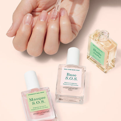 A hand applies Manucurist Rescue Kit nail oil with a dropper. Beside the hand are three treatments—Masque S.O.S., Base S.O.S., and Huile Verte—in clear glass bottles on a light pink surface.