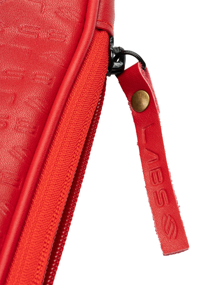 Red zippered pouch close-up showing zipper, brass-riveted leather pull tab with embossed letters