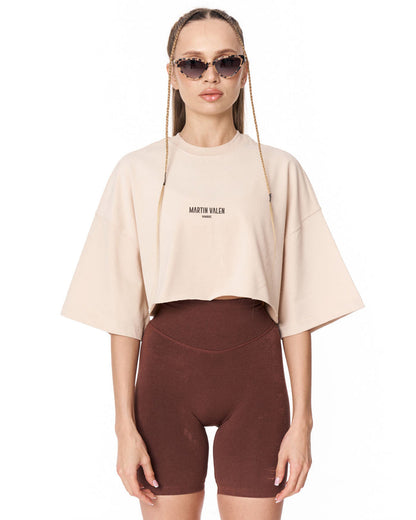 A model wearing a light beige cropped t-shirt with 'MARTIN VALEN' printed on the chest, paired with dark brown bike shorts, against a plain white background.