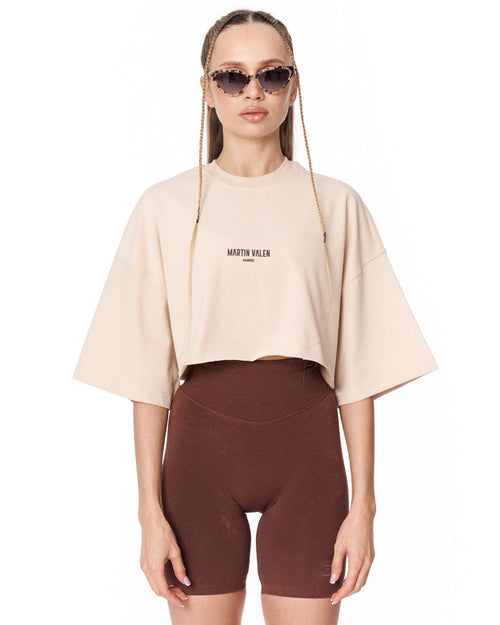 A model wearing a light beige cropped t-shirt with 'MARTIN VALEN' printed on the chest, paired with dark brown bike shorts, against a plain white background.