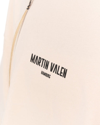 Close-up of a light beige or cream-colored garment with black printed text reading 'MARTIN VALEN' and a small portion of a zipper visible at the top left.
