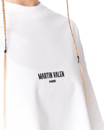 A close-up of a white crewneck sweatshirt featuring the black printed text 'MARTIN VALEN' and 'HAMBURG' on the chest, with braided drawstrings hanging down.