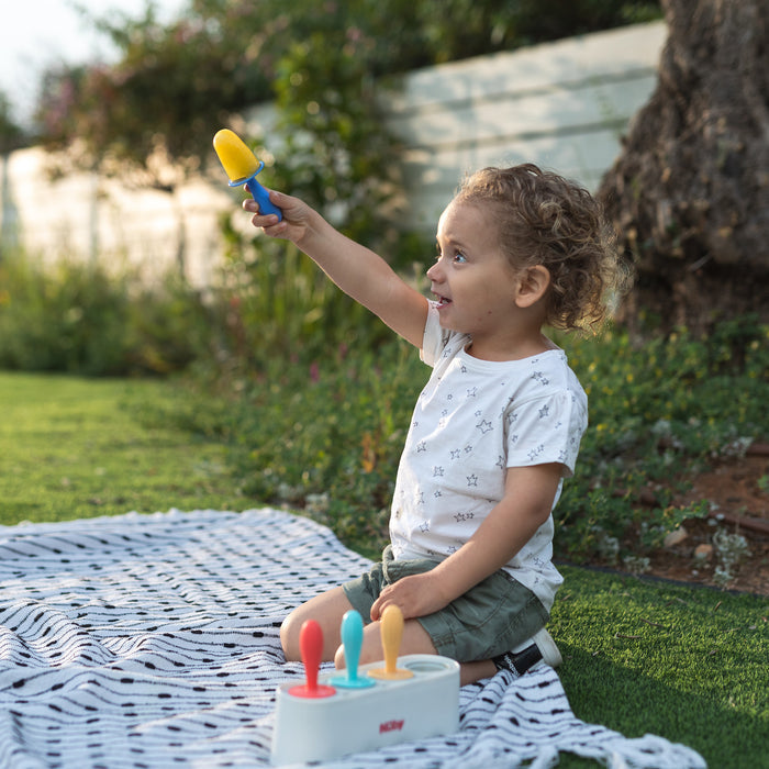 A young child with curly hair kneels on a blanket outdoors, smiling and holding up a yellow popsicle made with the Nuby Stick Pops Ice Pop Mold. The tray with three more colorful treats, ideal for teething babies, sits beside them amid greenery.