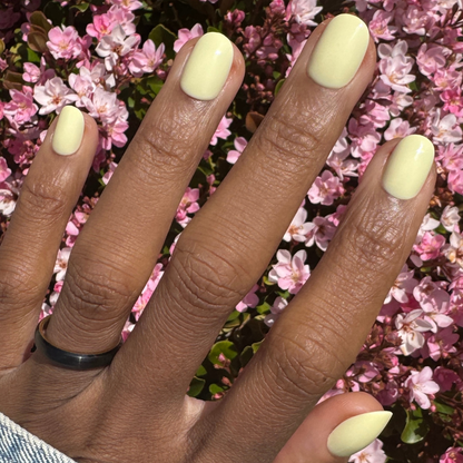 Sunny | Extra Short | Round - Tab Press-On nails in pastel yellow on hand outdoors against pink flowers in natural sunlight