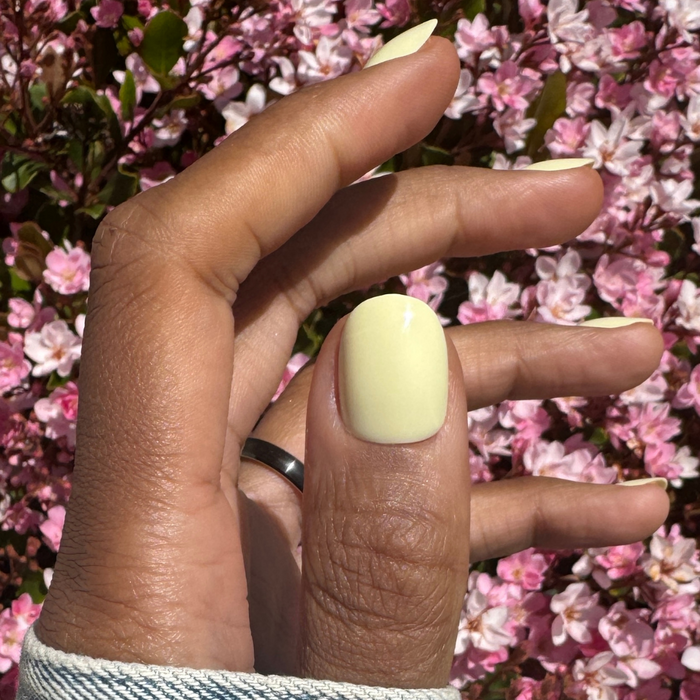 Sunny | Extra Short | Round - Tab Press-On nails in pastel yellow on natural hand outdoors against pink flowers in sunlight