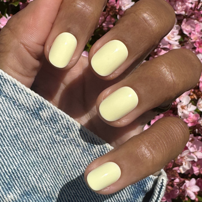 Sunny | Extra Short | Round - Tab Press-On nails in soft pastel yellow on hand outdoors with pink flowers and denim sleeve