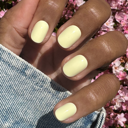 Sunny | Extra Short | Round - Tab Press-On nails in soft pastel yellow on hand outdoors with pink flowers and denim sleeve