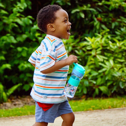 A young boy in a striped shirt and blue shorts smiles while running on a path, holding the Thirsty Kids REFLEX Easy Grip Water Bottle | Aqua Space. Lush green bushes are visible in the background.