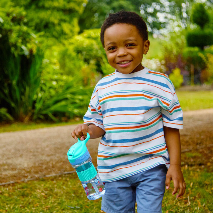 Smiling young boy in a striped shirt and blue shorts stands on a path, holding the Thirsty Kids REFLEX Easy Grip Water Bottle | Aqua Space. Lush green plants and trees fill the background.
