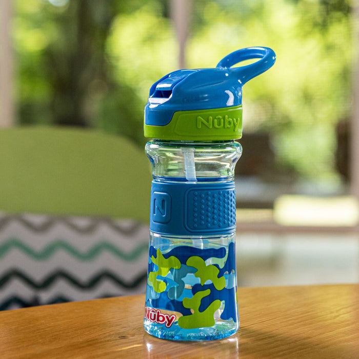 A Thirsty Kids REFLEX Easy Grip Water Bottle | Blue Camo rests on a wooden table, with blurred green foliage and indoor furniture in the background.