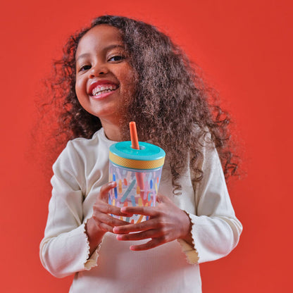 Smiling child with long curly hair holds a Thirsty Kids Super Sipper Straw Cup (2 Pack) | Aqua & Yellow, standing against an orange background.