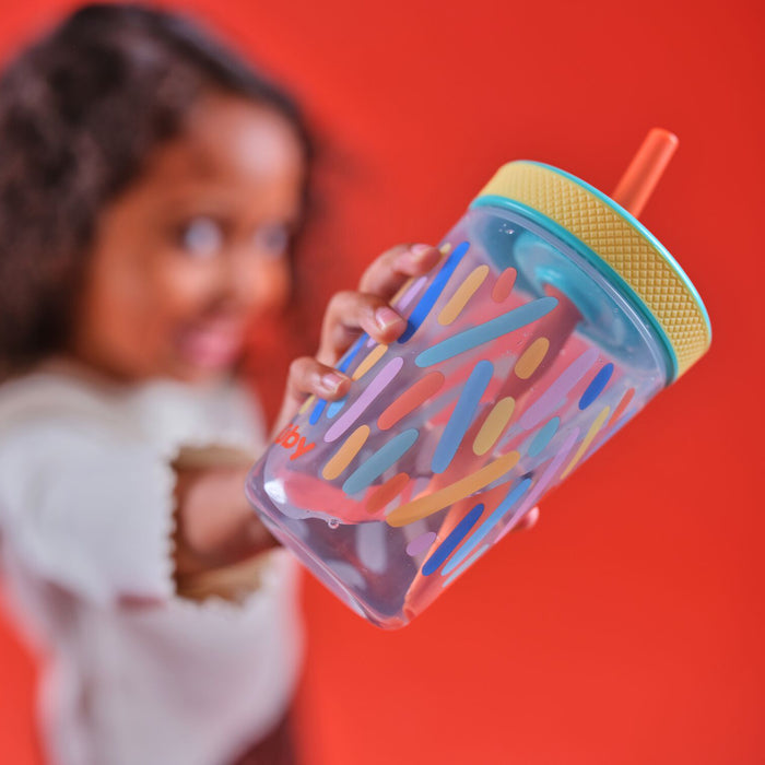 A child holds a Thirsty Kids Super Sipper Straw Cup (2 Pack) | Aqua & Yellow with playful shapes, yellow lid, and orange straw against a bright red background. The smiling child appears out of focus.